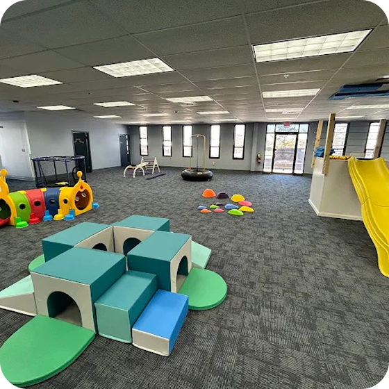 Indoor playroom featuring soft play foam blocks, a colorful caterpillar tunnel, balance pods, and a yellow slide.