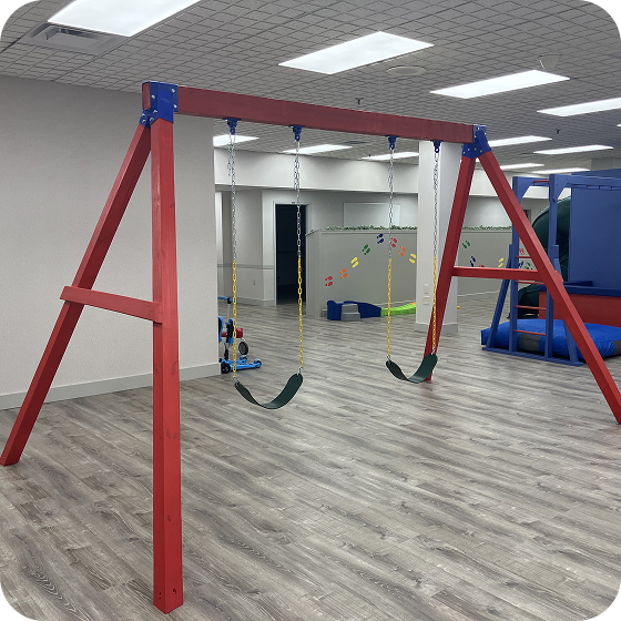 Indoor red wooden swing set with two swings in a large playroom.