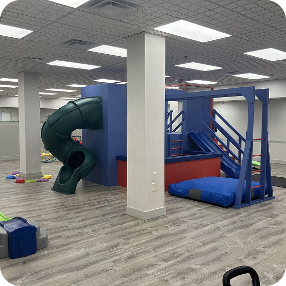 Indoor playground room featuring a green curved slide, climbing structure, stairs, and a blue safety mat.