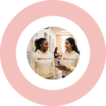 Two female staff members in matching OnTarget ABA hoodies collaborating at a table with learning materials and a tablet.